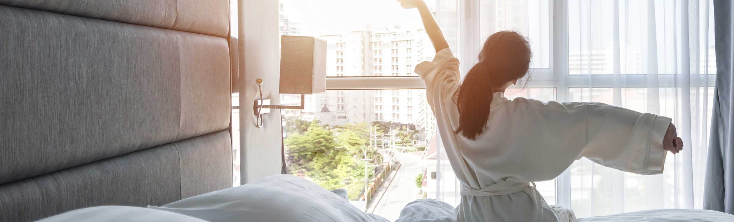 Woman in a robe sitting on a bed facing a sunny window whole stretching her arms