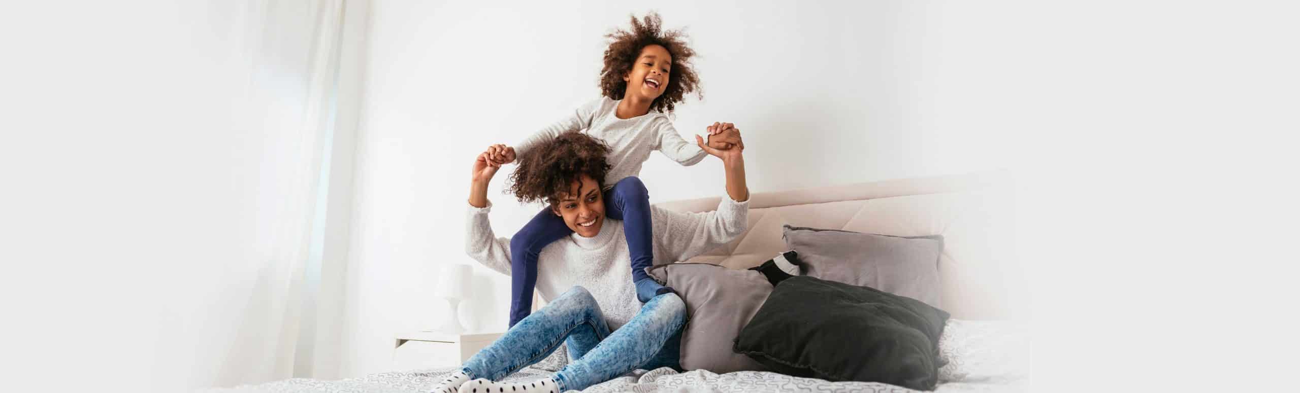 Mother smiling while sitting on a bed with her daughter on her shoulders.