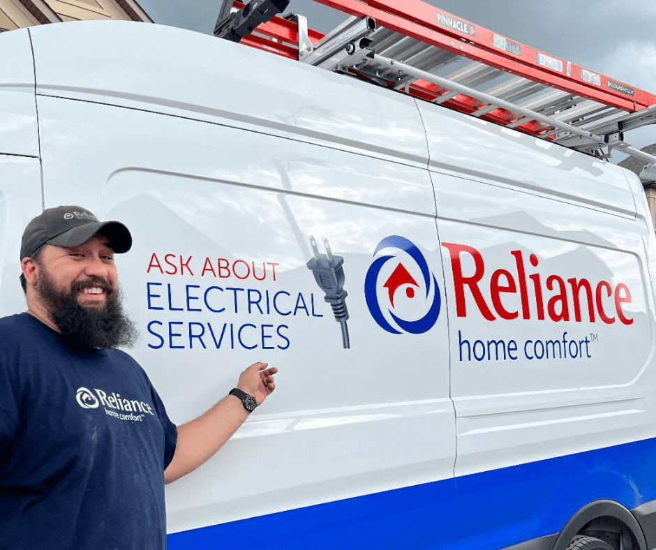 A Reliance Electrician stands beside a Reliance truck pointing to the words, Ask Us About Electrical Services